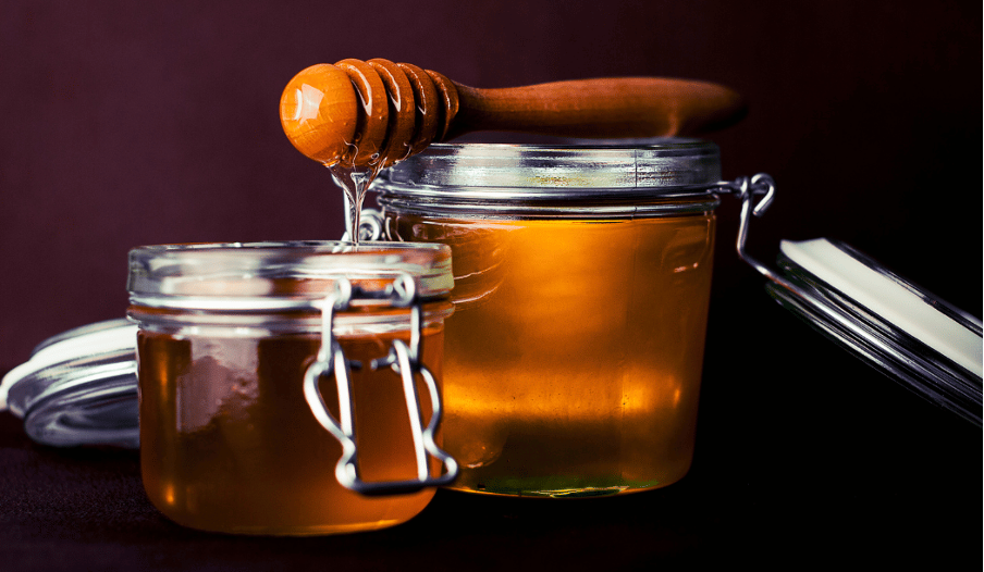 Es sind zwei Honiggläser mit klaren bzw. flüssigem Honig und einem Honiglöffel zu sehen.